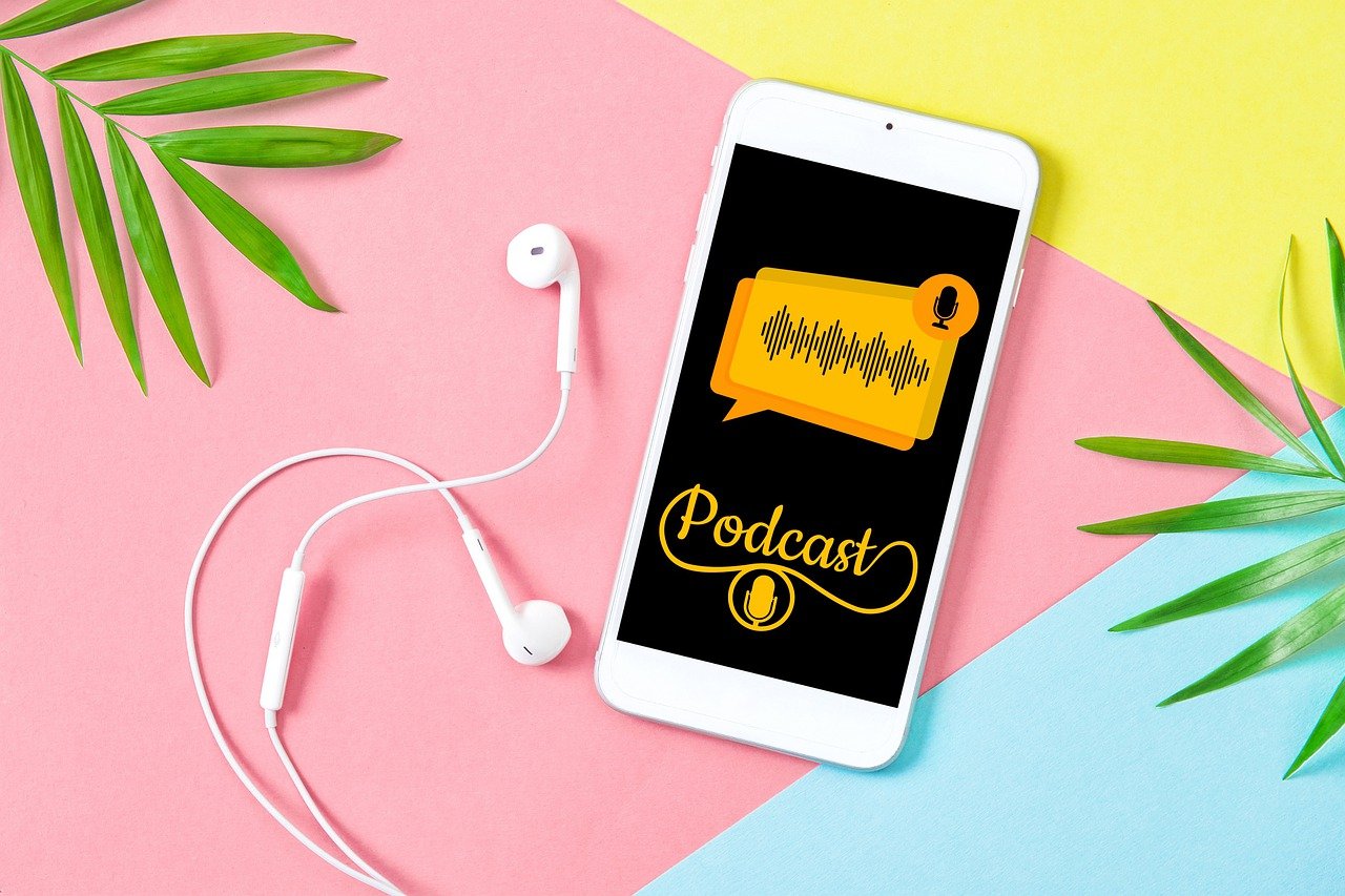 mobile phone with white earbuds on a colorful pastel (pink, yellow, blue) and green leaves on left and right side. Screen on phone shows the word "Podcast" in a yellow chat box.